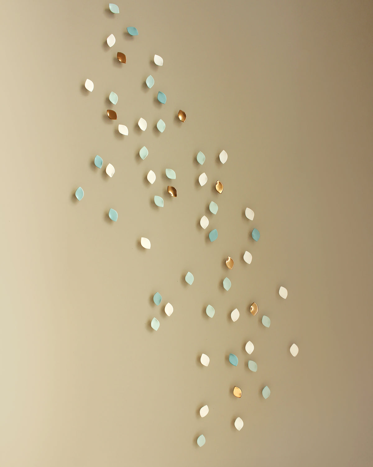 Decorative wall art sculptures shaped like small leaves or petals, scattered in a diagonal cluster on a warm beige wall. The artwork comes in three colors: white, light blue, and metallic gold.