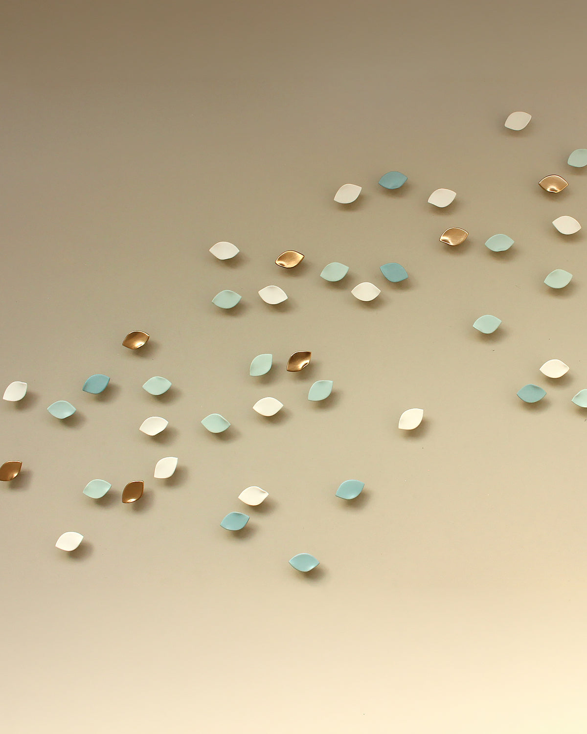 Decorative wall sculpture in the shape of small leaves, scattered diagonally on a warm beige background. The petals come in colours mid teal blue, light teal blue, metallic gold, and white.