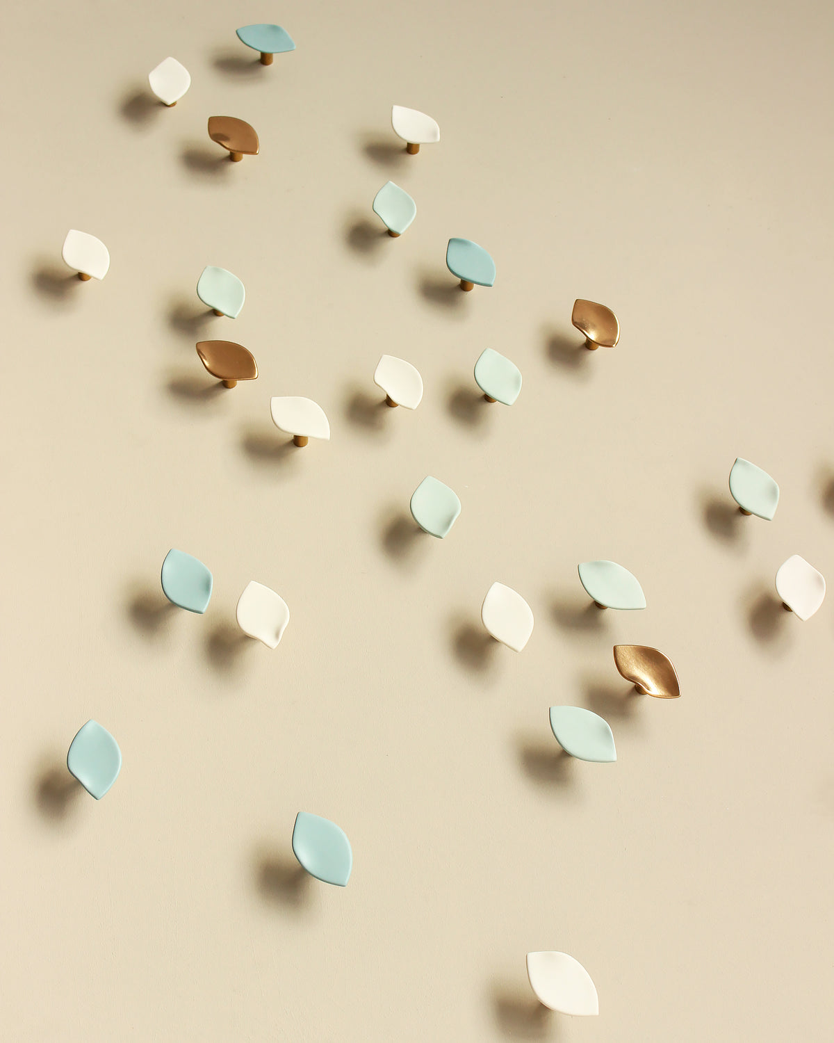 A collection of small, leaf-shaped wall sculptures in light blue, white, and metallic gold, scattered across a warm beige background in a flowing arrangement.