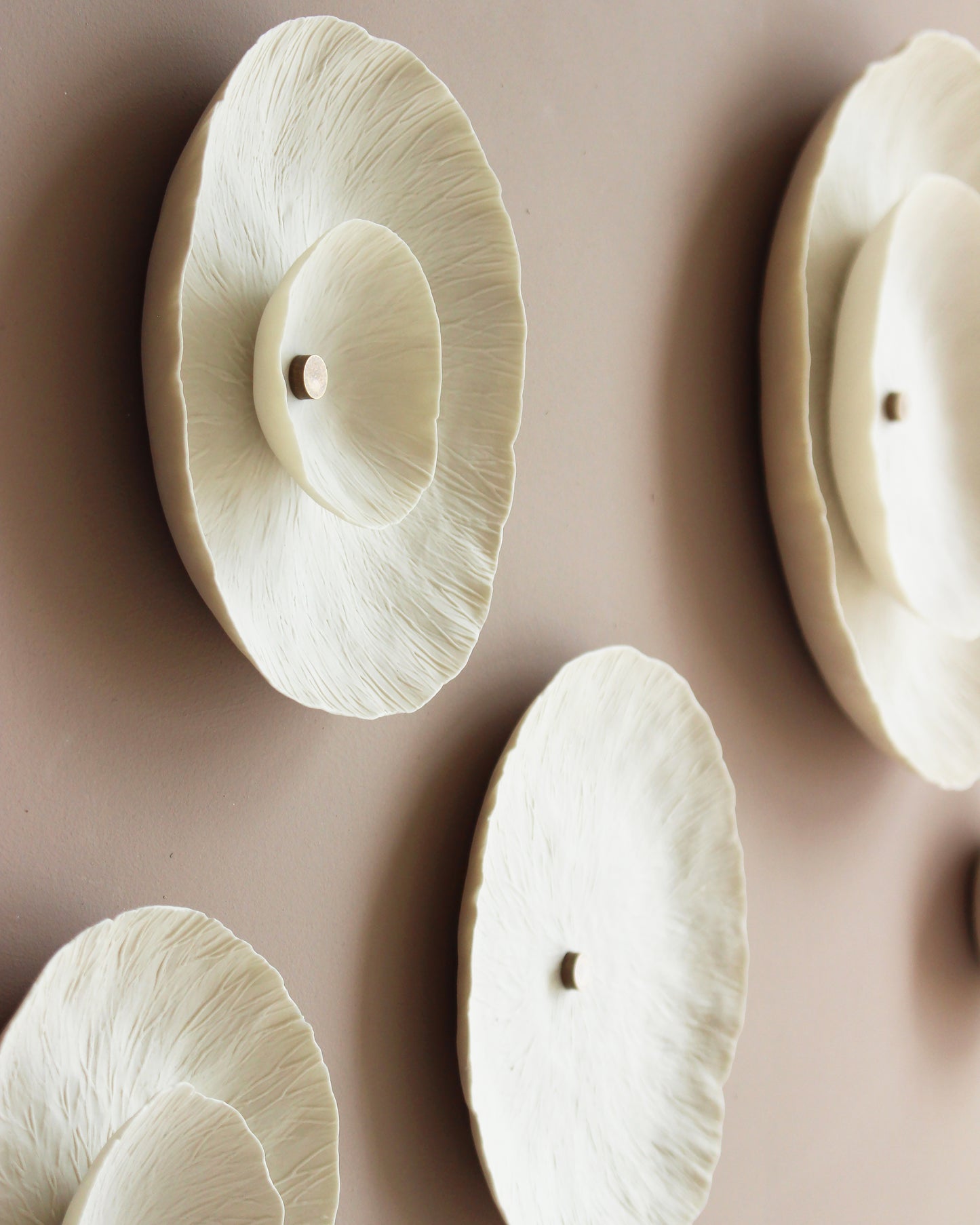 A close-up of a sculptural circular shaped wall decor piece in off white/cream natural porcelain colour, mounted on a warm taupe wall. The design features layered, textured patterns, similar to lichen and a small golden stud in the centre.
