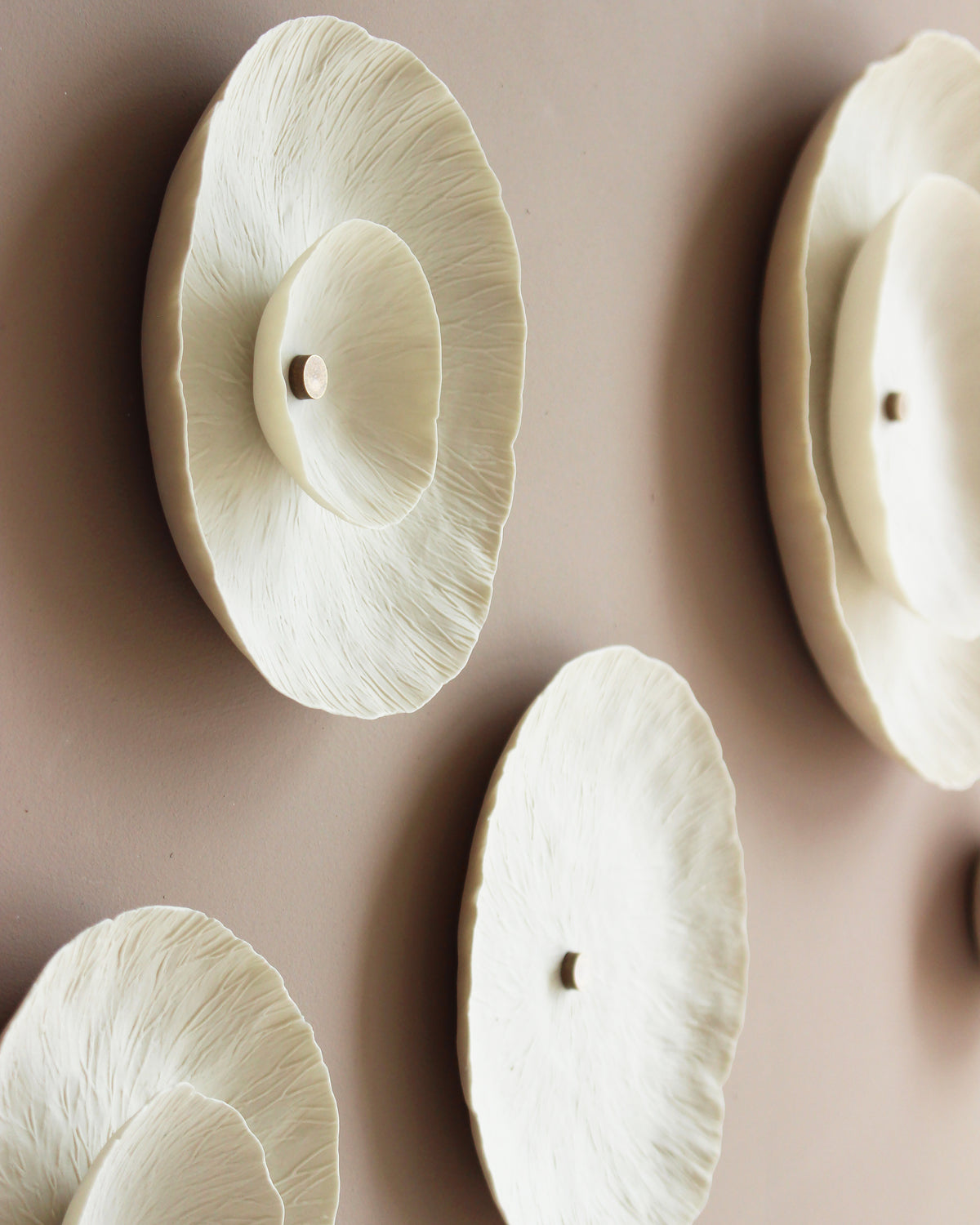 A close-up of a sculptural circular shaped wall decor piece in off white/cream natural porcelain colour, mounted on a warm taupe wall. The design features layered, textured patterns, similar to lichen and a small golden stud in the centre.