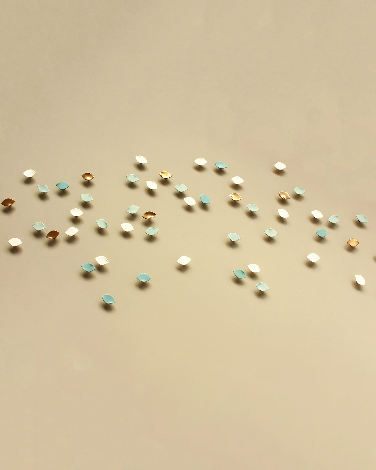 Small scattered leaf shaped wall sculptures in white, light blue, and metallic gold on a plain beige background.